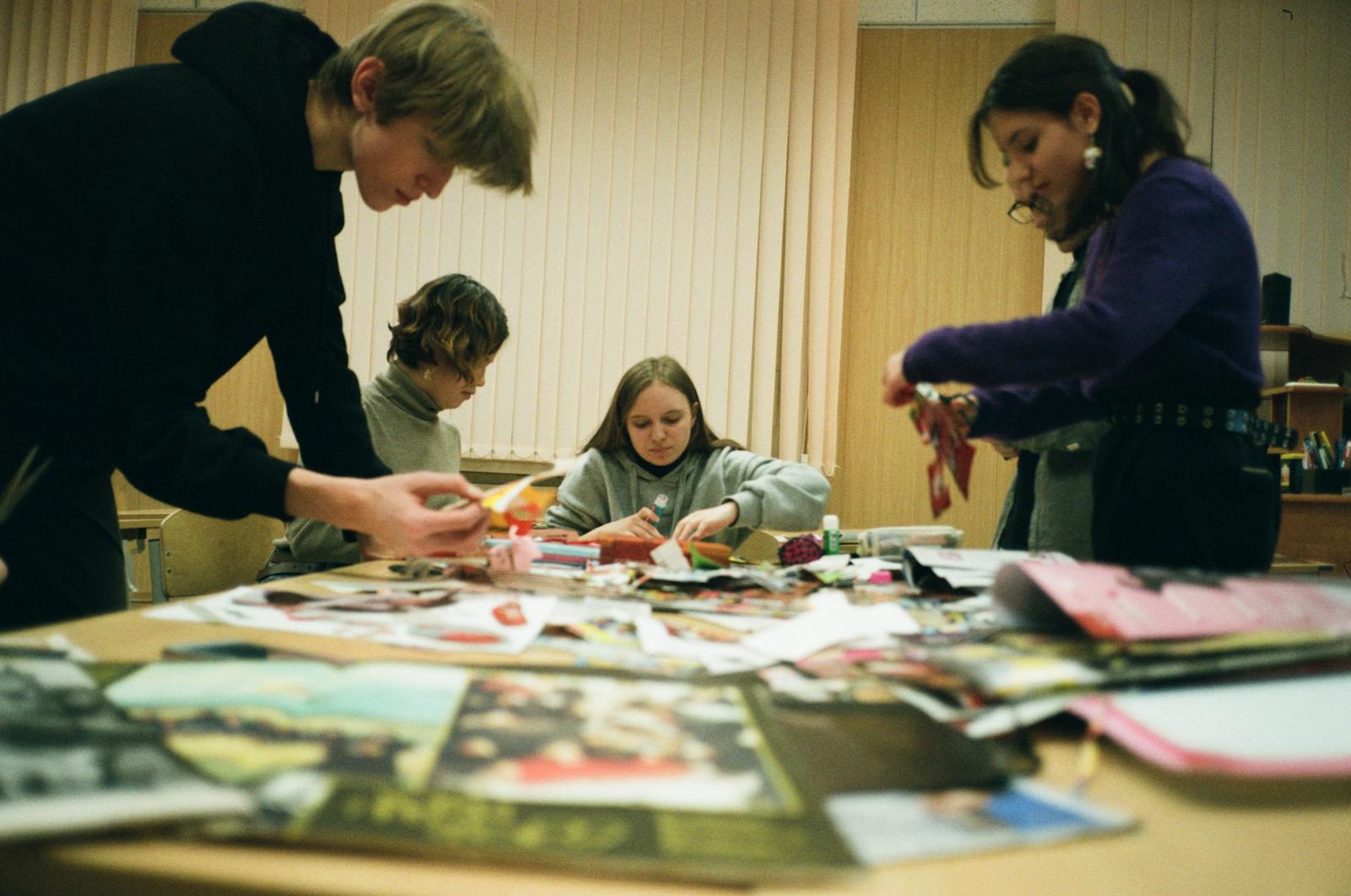 Adolescents cutting images with scissors from bright magazines while sitting at table in classroom