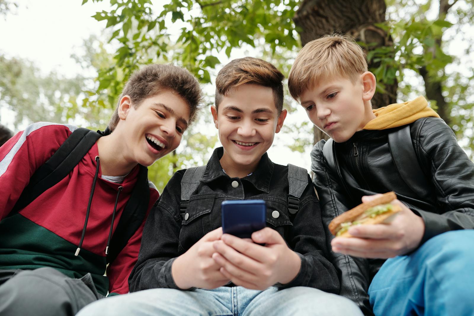 Three teenage boys sitting outdoors enjoying and laughing while using a smartphone.