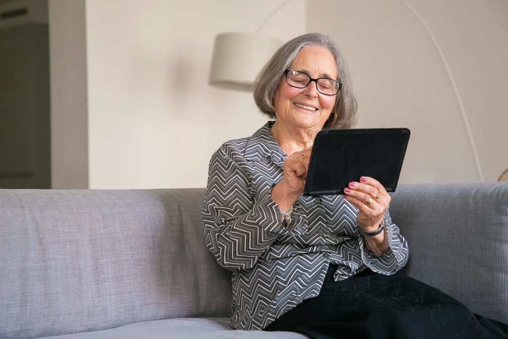 Elderly caucasian woman enjoying a tablet indoors on a comfortable sofa.