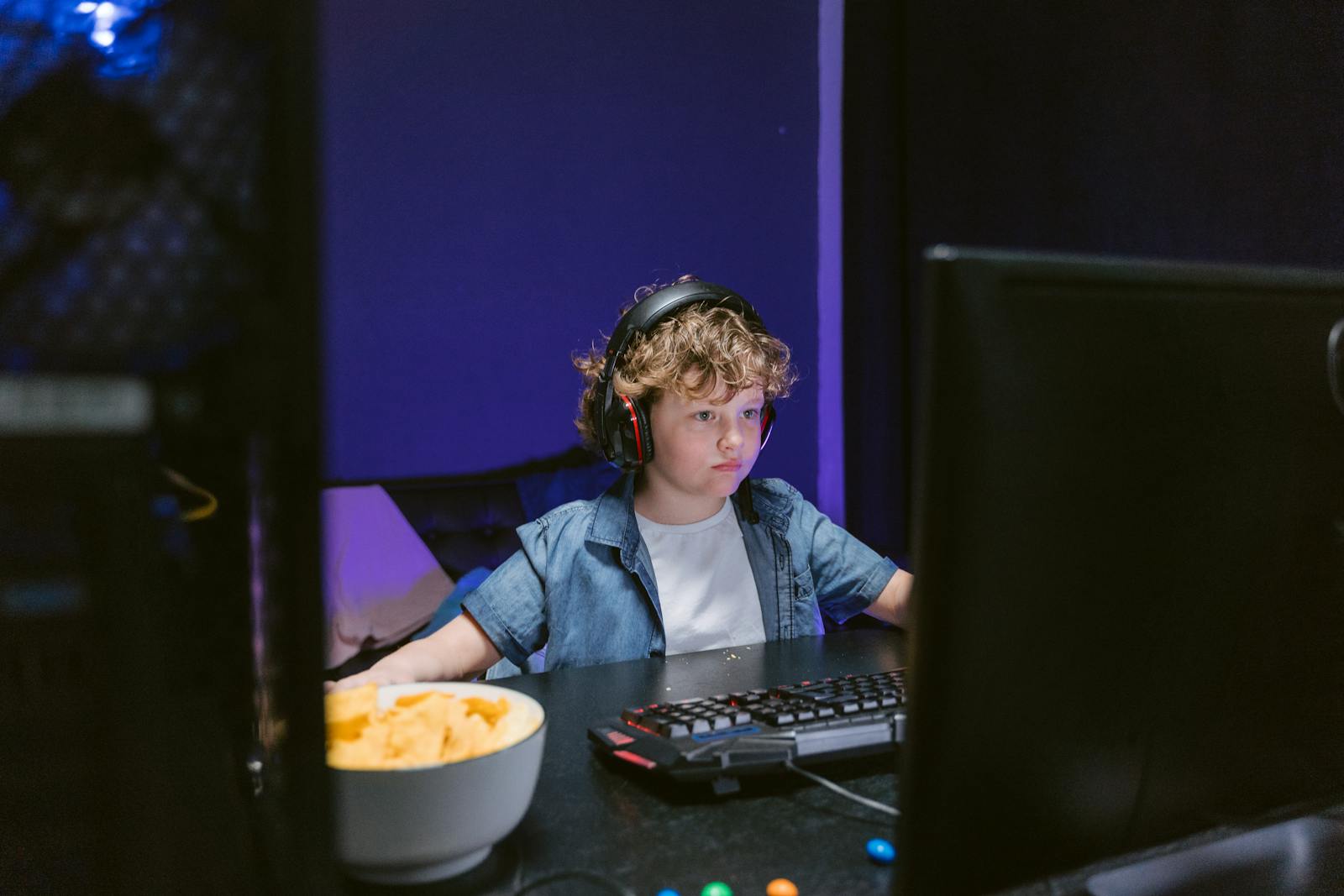 A child wearing headphones intensely playing video games in a dimly lit room with snacks nearby.