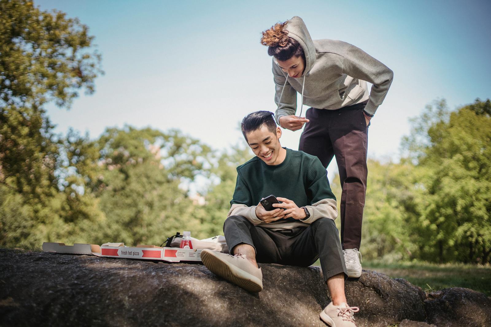 Two friends enjoying pizza and browsing smartphone in a park setting.