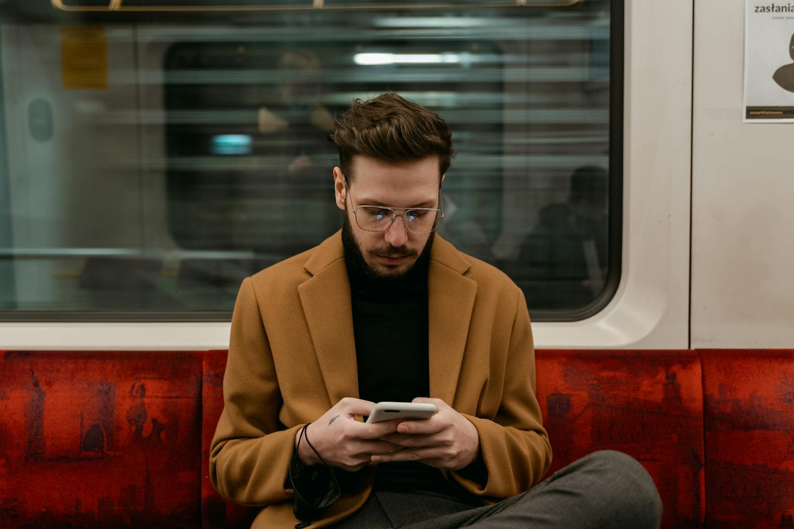 A man in a brown coat uses his smartphone while commuting on a train. Urban city life captured indoors.